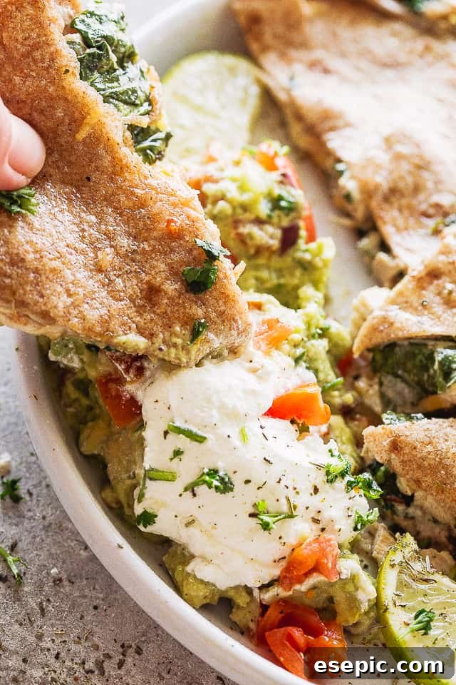 These baked chicken quesadillas are stuffed with fresh spinach, melted cheese and shredded chicken. It's 100% kid approved and oh so good! A hand dipping a half-quesadilla into a bowl of sour cream and guacamole.
