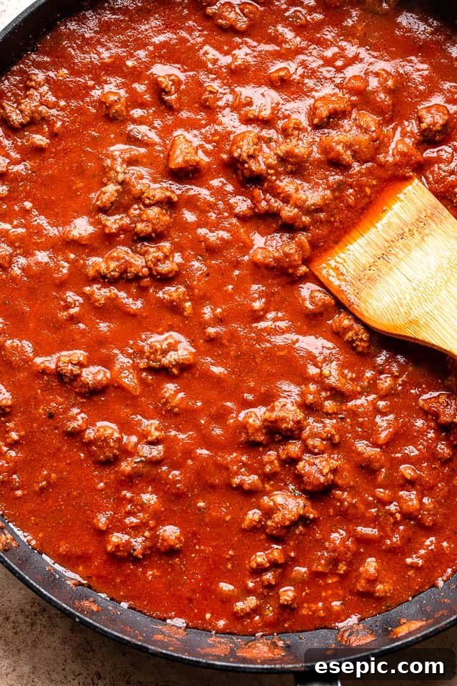 The Ultimate Cheesy Baked Ziti 3 Cooking ground beef, onions, and garlic with spices in a large black skillet before adding tomato sauce.