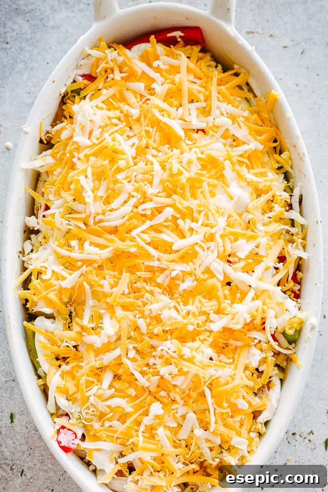 Shredded cheese sprinkled generously over chicken and peppers in a white oval baking dish, before baking.
