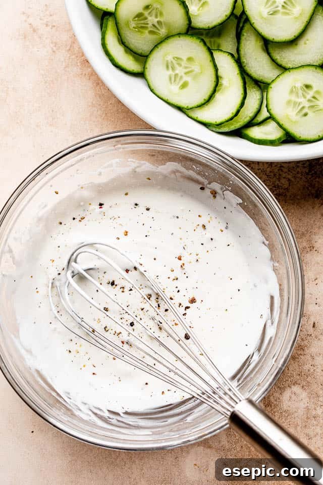 A glass bowl holds the smooth, white sour cream dressing, with a whisk resting inside, ready for mixing.