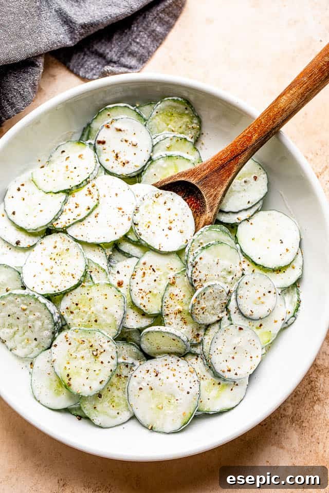 A wooden spoon gently mixes thinly sliced cucumbers coated in a creamy sour cream dressing in a white salad bowl.