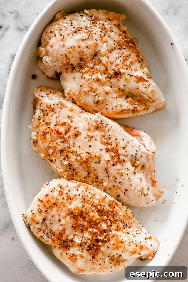 Top-down view of four seasoned boneless, skinless chicken breasts arranged neatly in a white oval baking dish, ready for dressing and cheese.