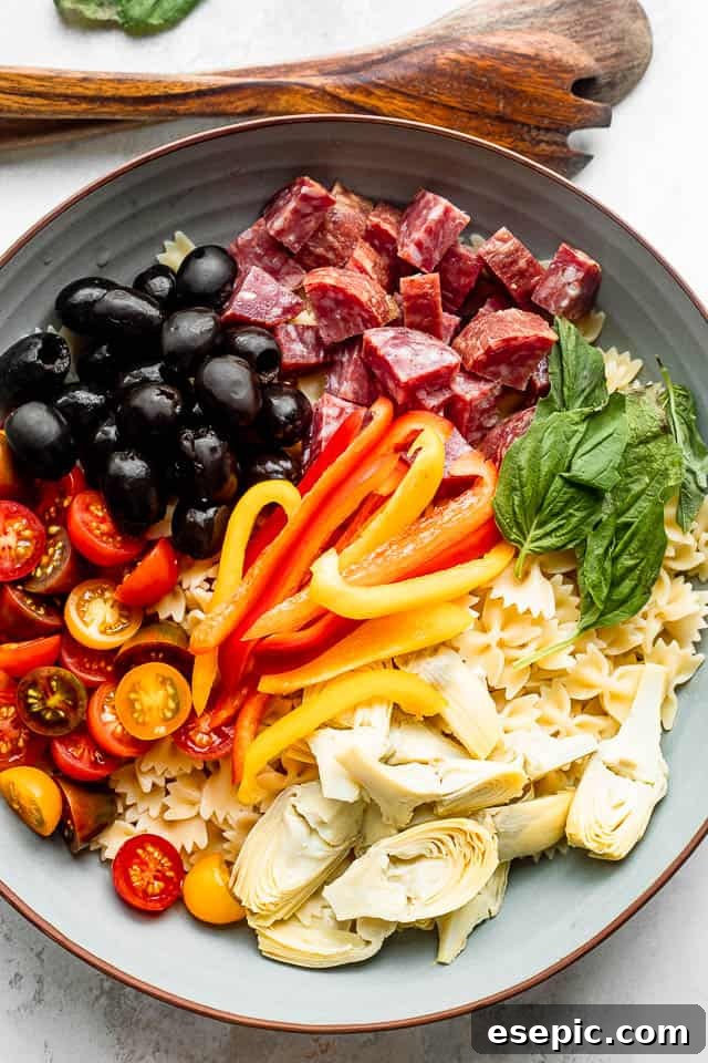 Artistically arranged ingredients for Italian Antipasto Pasta Salad, including pasta, olives, salami, cheese, artichokes, tomatoes, basil, and peppers on a wooden surface next to a bowl.
