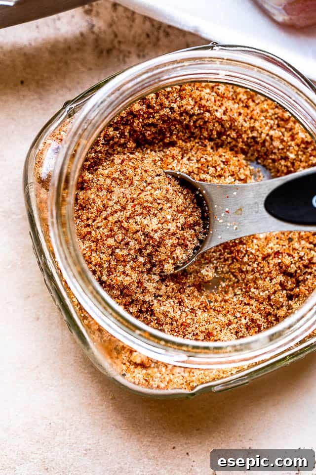 A silver tablespoon filled with a savory brown sugar seasoning rub in a glass jar