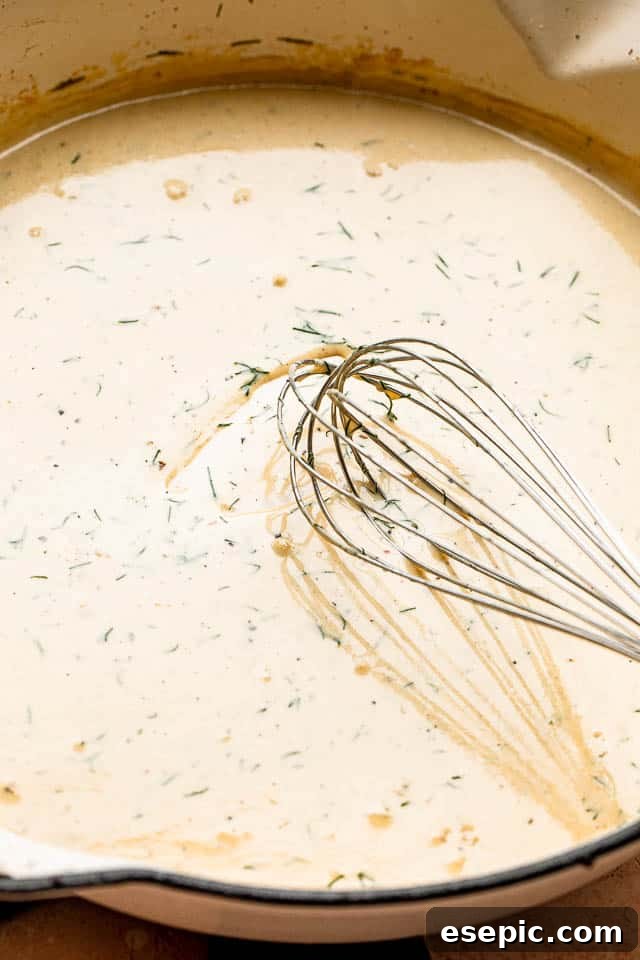 A close-up shot of a whisk actively mixing a creamy, light yellow sauce in a white skillet, illustrating the smooth consistency of the dill sauce.