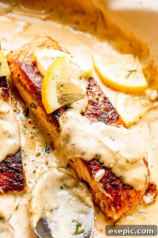 Close-up of a single cooked pan-seared salmon fillet, elegantly topped with a generous spoon of creamy dill sauce and a thin slice of lemon, presented on a white plate.