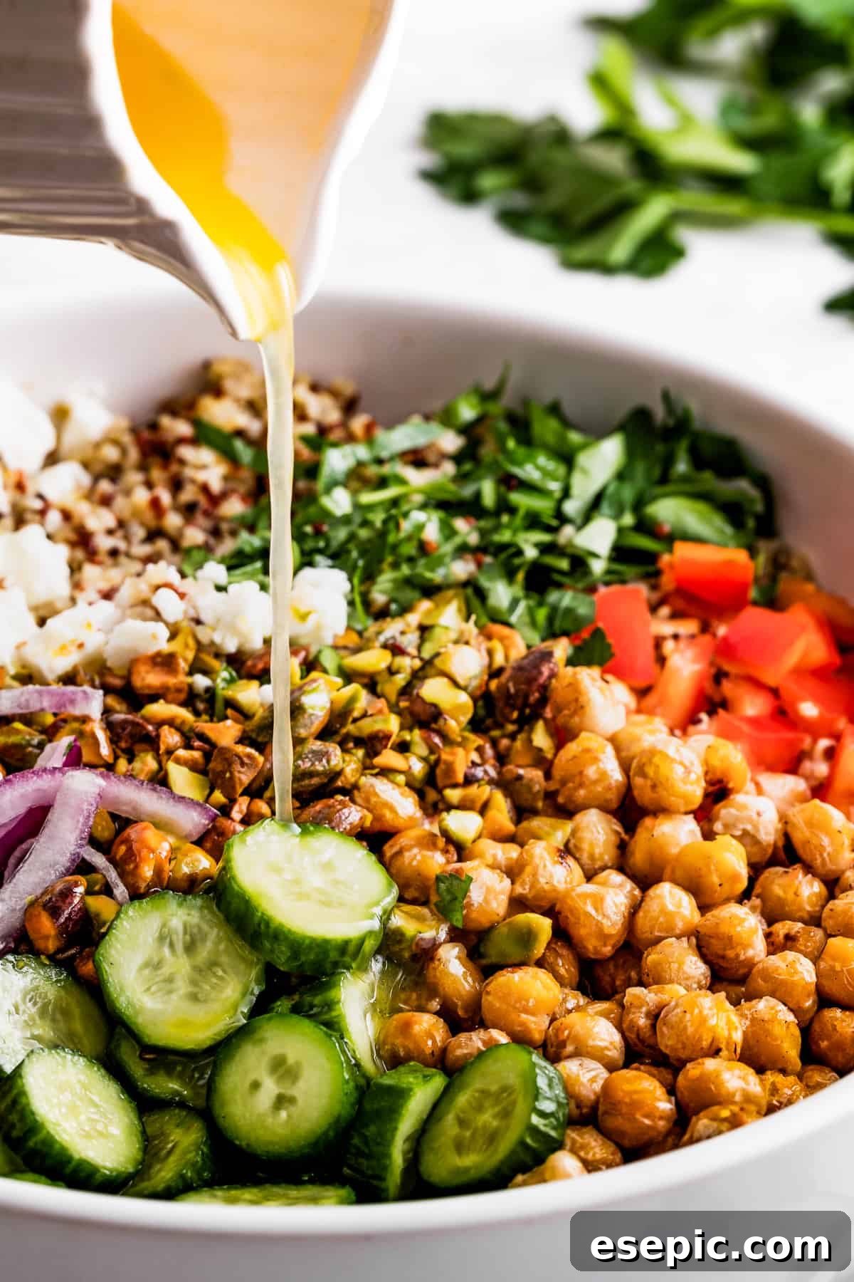 Close-up shot of homemade dressing being poured over the vibrant Jennifer Aniston salad ingredients, including chickpeas, sliced cucumbers, red onions, bell peppers, fresh herbs, and crumbled feta cheese.