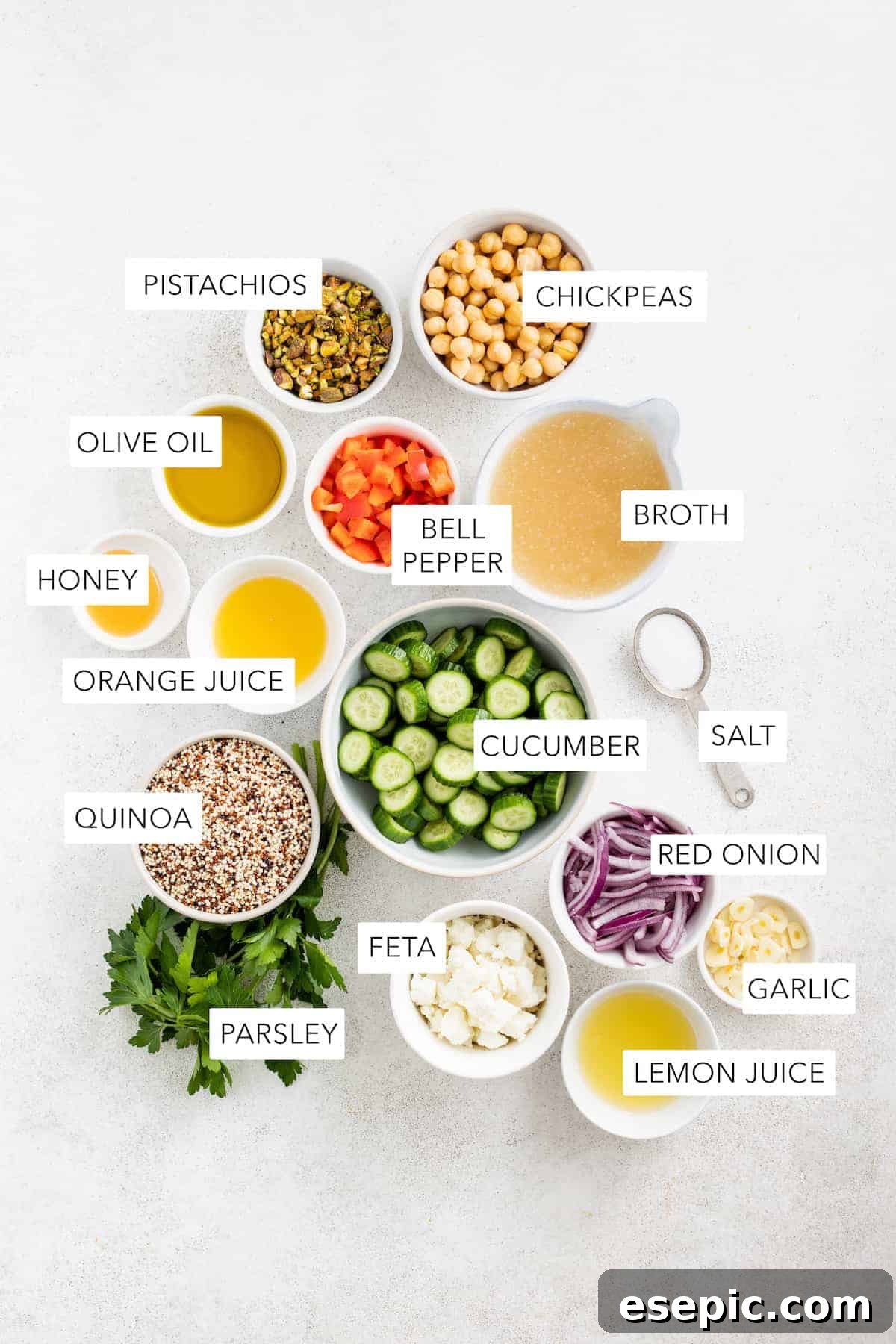 A collection of fresh, wholesome ingredients laid out for the Jennifer Aniston Quinoa Salad, including cucumbers, bell peppers, red onion, quinoa, chickpeas, feta, pistachios, and various dressing components.