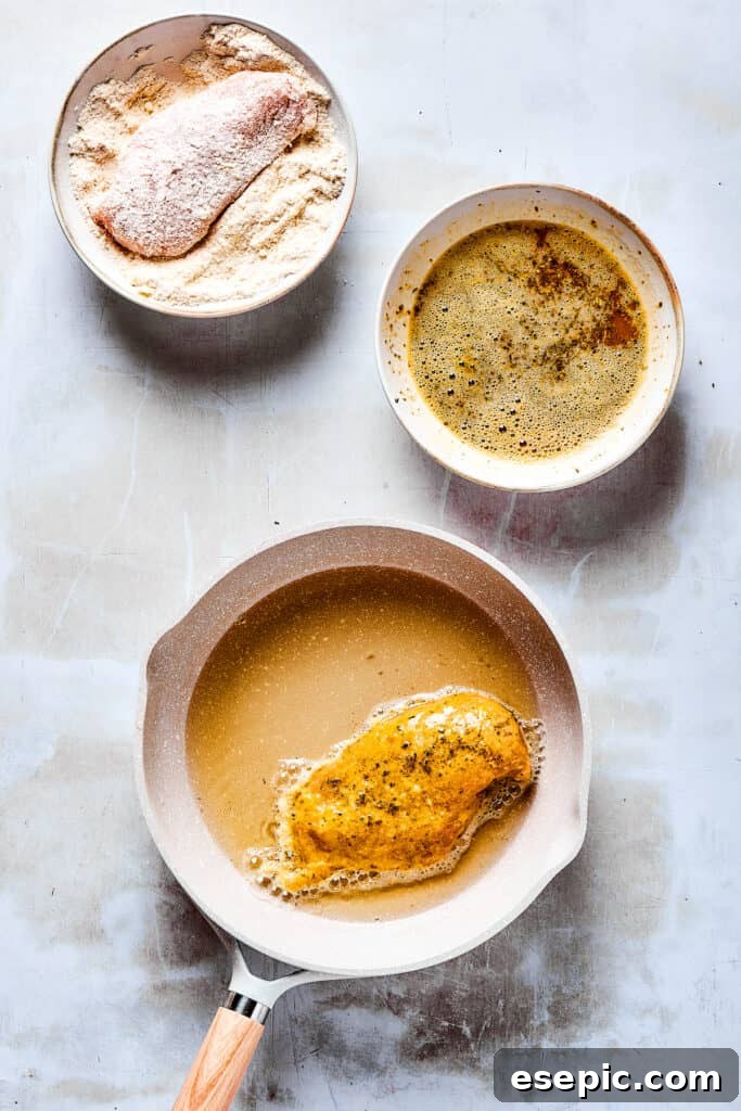 A chicken breast in a dish of dry breading ingredients, next to a dish of wet breading ingredients, and a skillet with one breaded chicken breast cooking in oil.