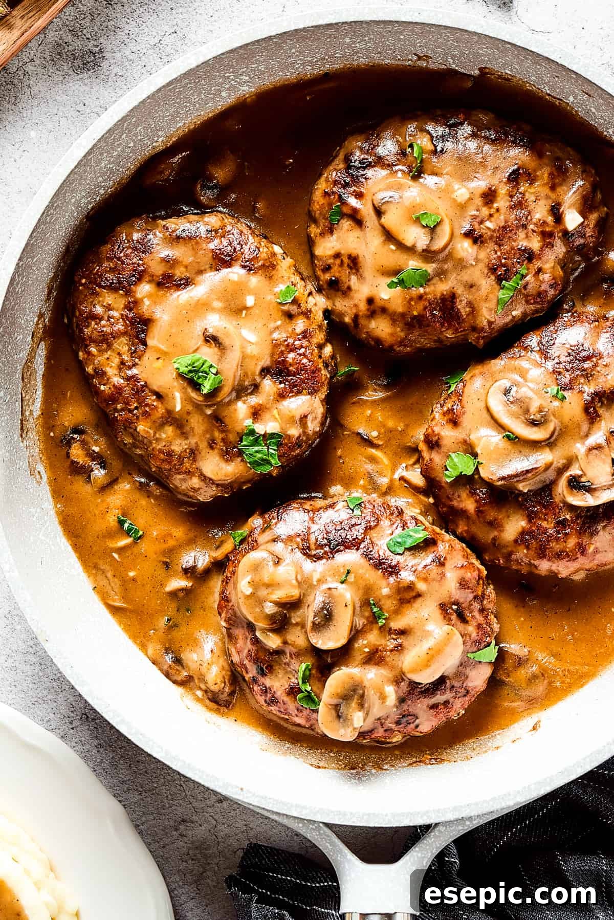 Four golden-brown Salisbury steaks nestled in a rich mushroom gravy in a cast-iron skillet, surrounded by fresh herbs and a cloth napkin.