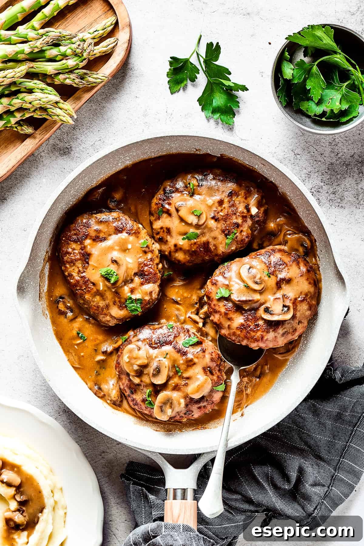 A vibrant skillet of Salisbury steaks in gravy, garnished with fresh herbs and surrounded by colorful vegetables and a rustic cloth.