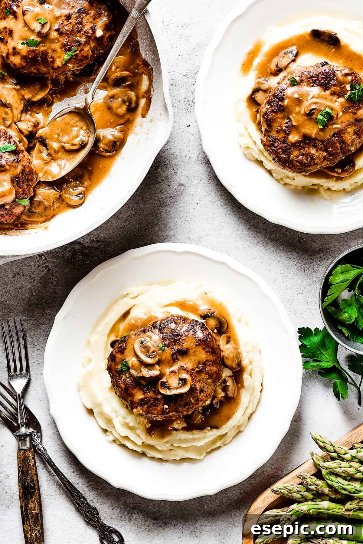 A skillet of Salisbury steaks in gravy next to two dinner plates, each with a serving of Salisbury steak and mashed potatoes.
