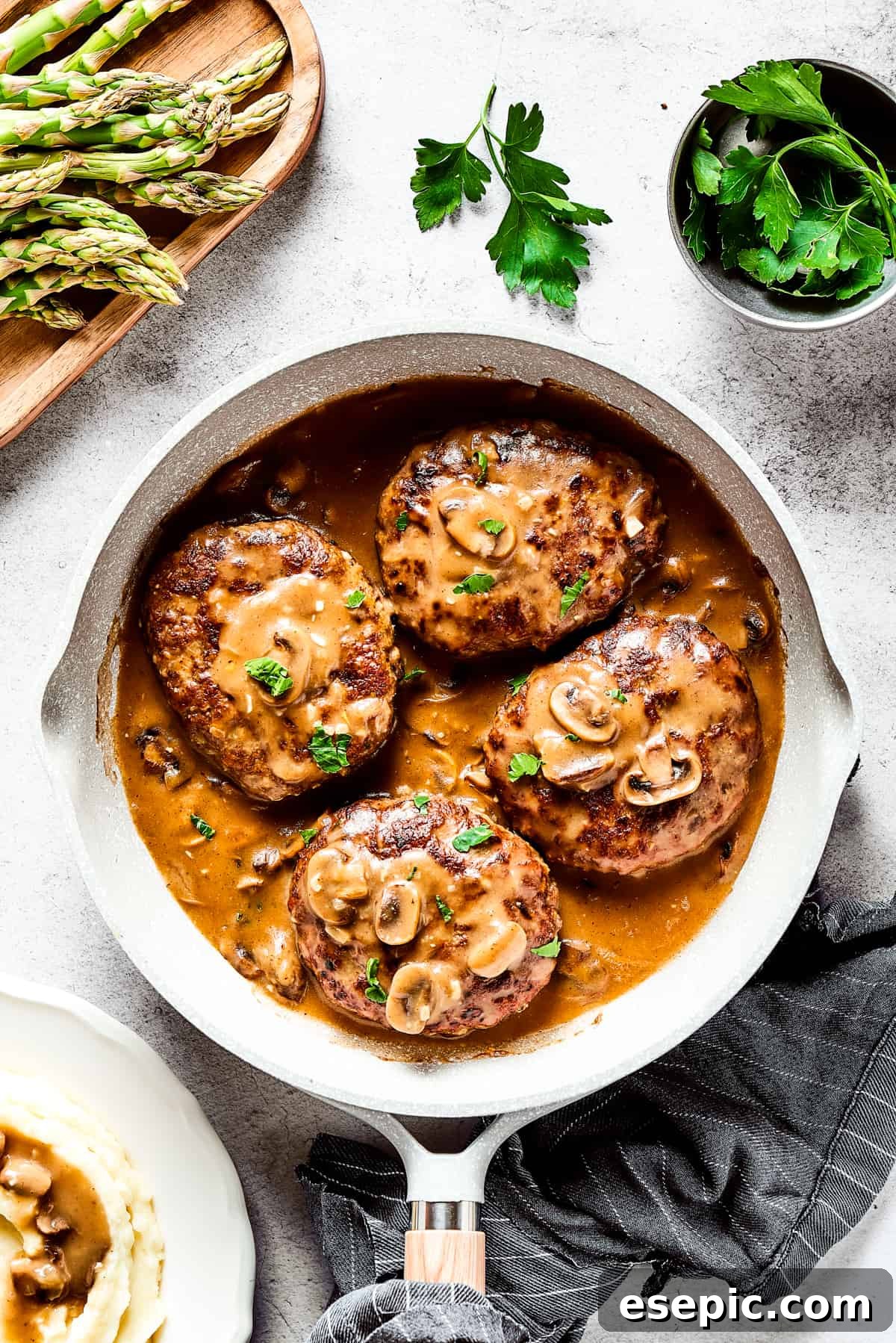 An overhead shot of four Salisbury steaks in a skillet, swimming in thick mushroom gravy, with fresh garnishes.