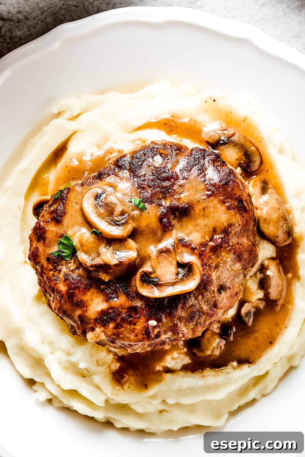A serving of Salisbury steak with mushroom gravy over mashed potatoes on a white dinner plate, garnished with fresh parsley.