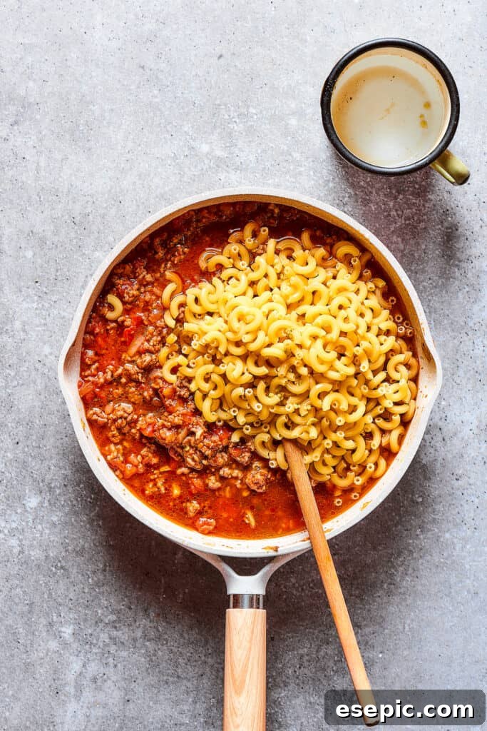 Adding uncooked elbow macaroni pasta directly into the rich beef and tomato sauce in the skillet.