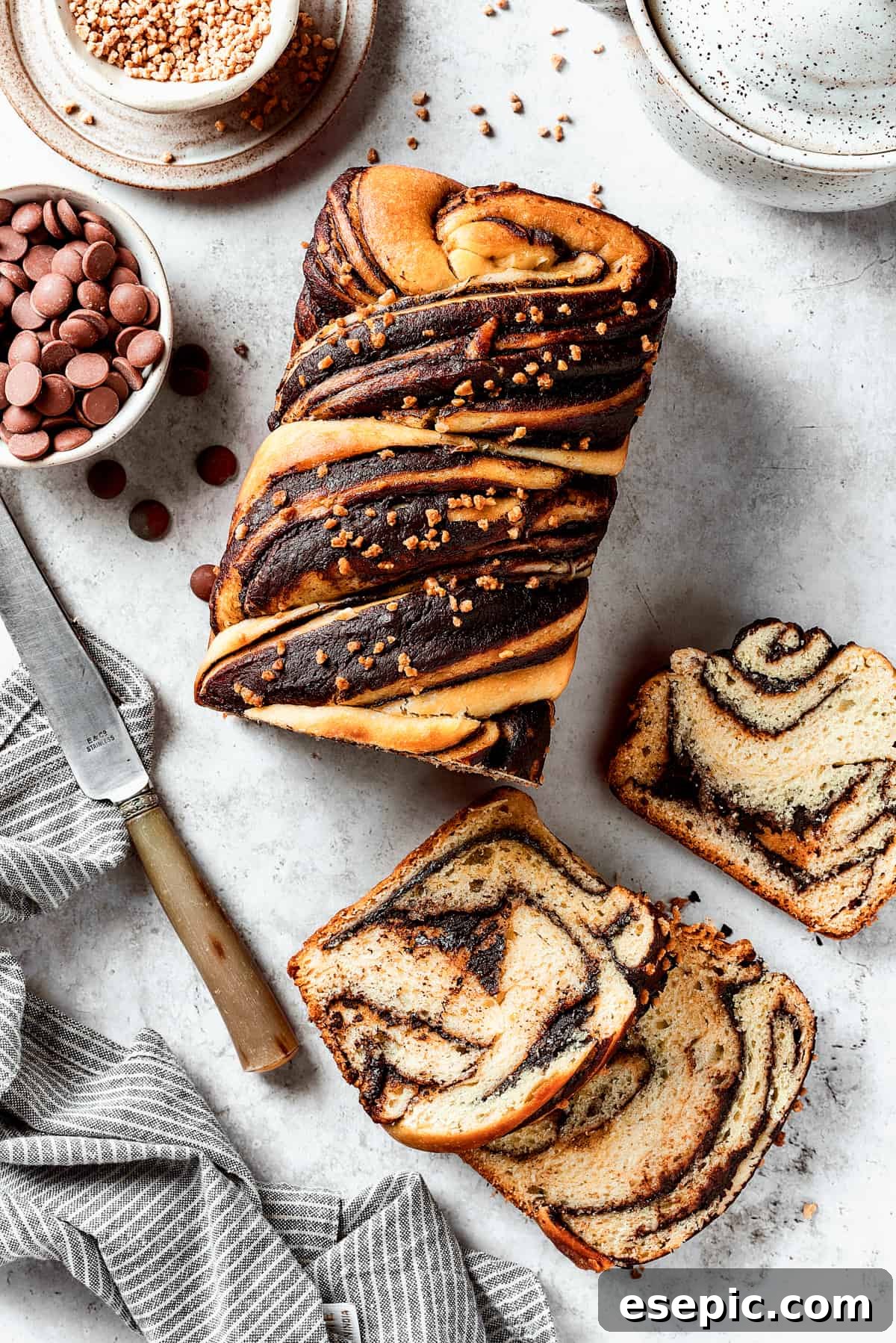 Sweet, puffy Chocolate Babka is an old-fashioned yeast bread swirled with layers of chocolate. Perfect for a special breakfast or dessert. A golden-brown chocolate babka loaf, partially sliced, with slices arranged alongside a bread knife and a bowl of chocolate chips.