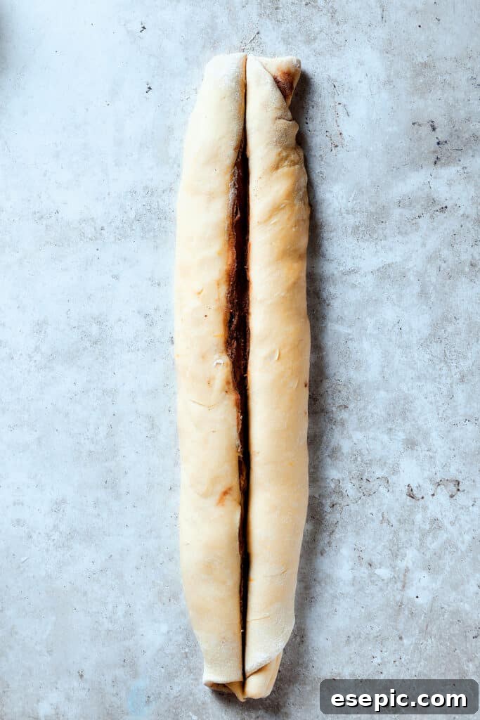 Sweet, puffy Chocolate Babka is an old-fashioned yeast bread swirled with layers of chocolate. Perfect for a special breakfast or dessert. A knife carefully slicing a roll of dough lengthwise, revealing the chocolate layers within.