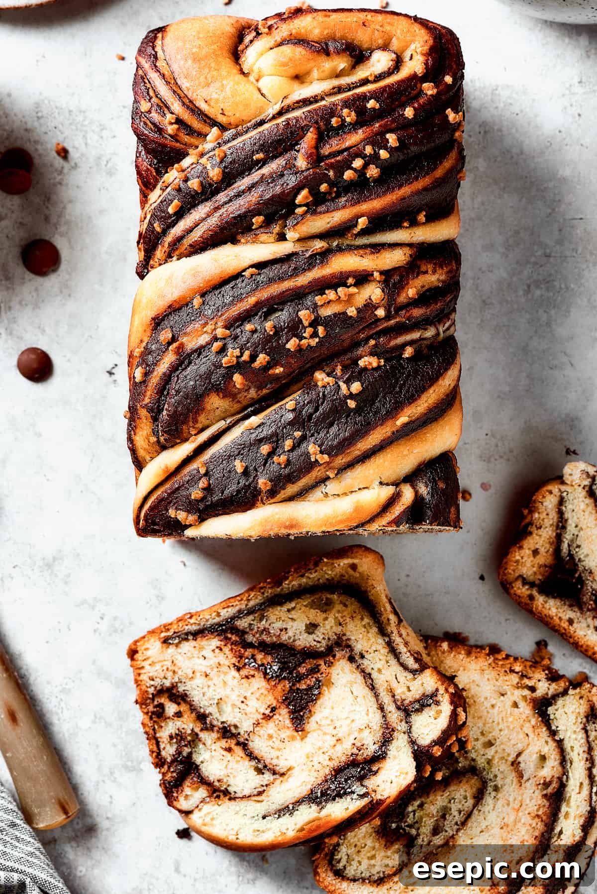 Chocolate Babka - Traditional Chocolate Swirl Bread! A loaf of chocolate babka that has been partially sliced. The slices are laid out on the work surface, along with a bread knife and a dish of chocolate chips.
