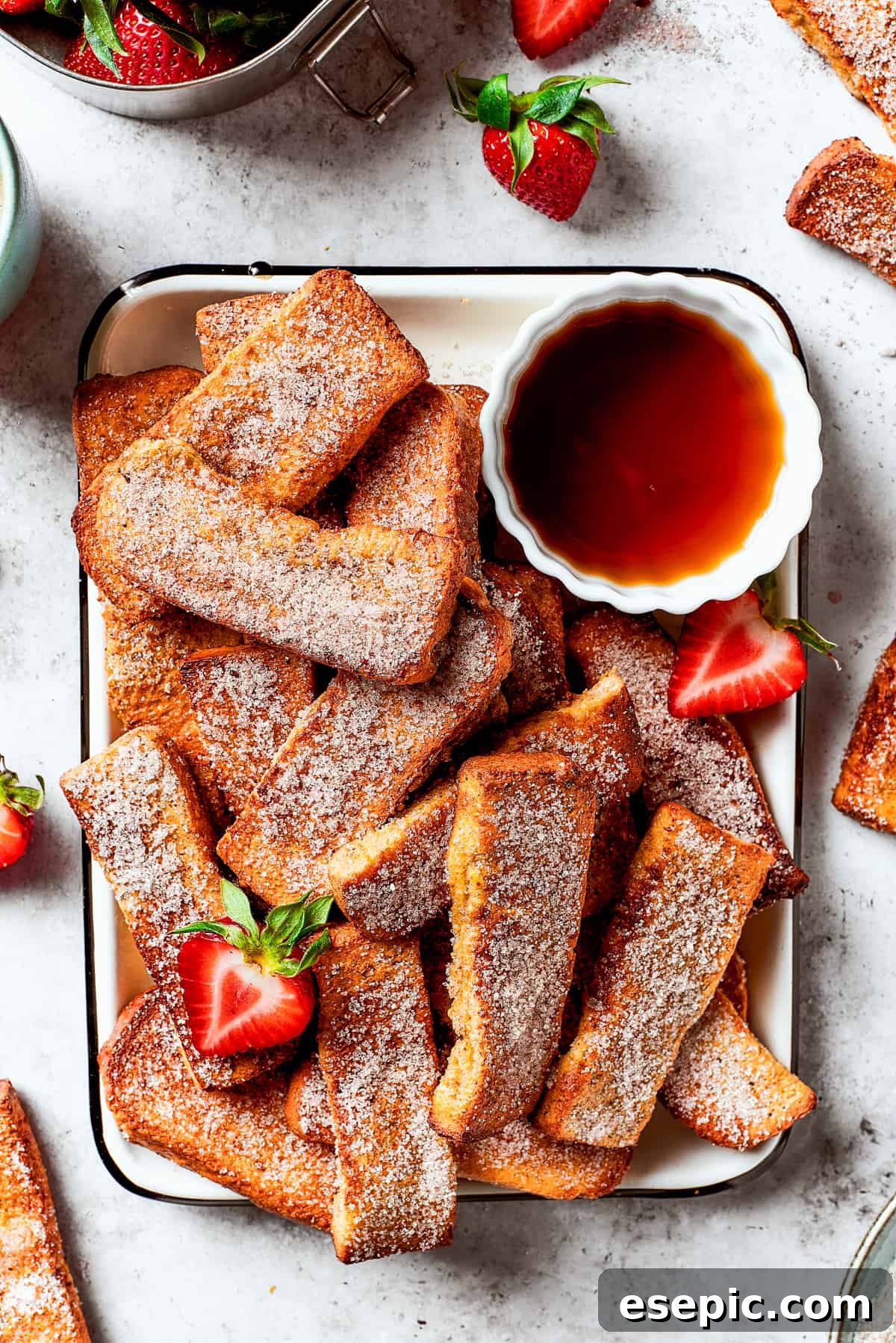 Air fryer French toast sticks on an enamel tray with a cup of maple syrup and fresh strawberries.