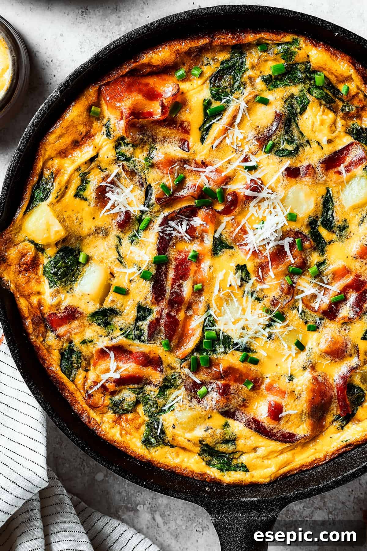 Spinach, Bacon, and Potato Frittata is a healthy, farmhouse-style dinner that's easy to make, and loaded with rich flavor. Perfect easy meal! Close-up overhead shot of a fully-cooked frittata in a cast-iron pan, showcasing its golden-brown top and vibrant fillings.
