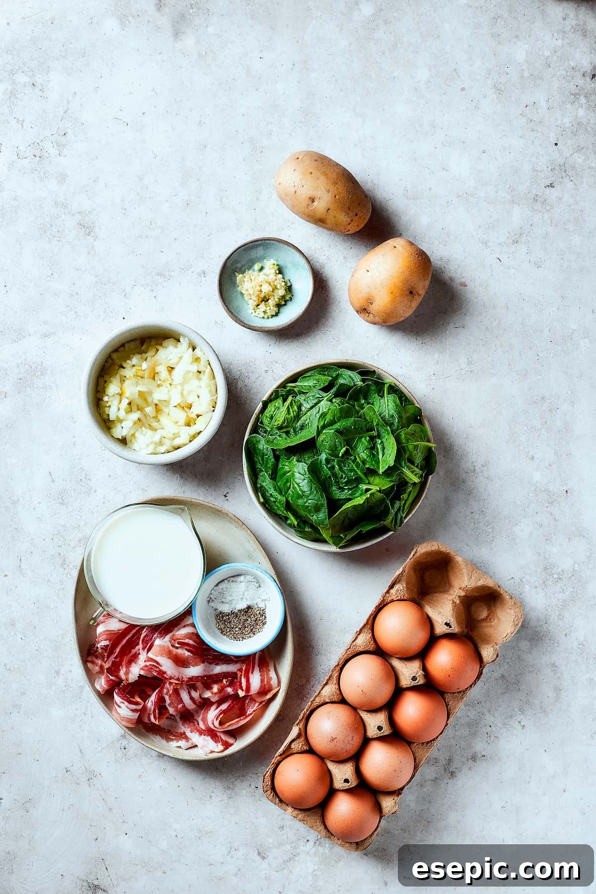 Spinach, Bacon, and Potato Frittata is a healthy, farmhouse-style dinner that's easy to make, and loaded with rich flavor. Perfect easy meal! From top: Potatoes, fresh spinach, whisked eggs, crispy bacon, salt and pepper shakers, a jug of milk, diced onion, and minced garlic laid out on a wooden surface.