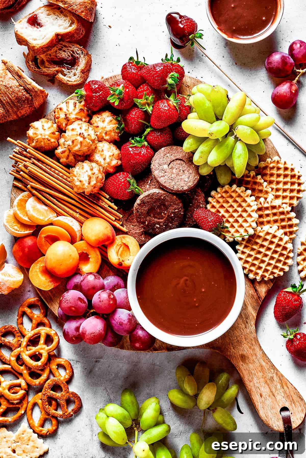 An inviting dessert board featuring a central bowl of rich chocolate fondue, surrounded by various fruits, waffles, and pretzels for dipping.
