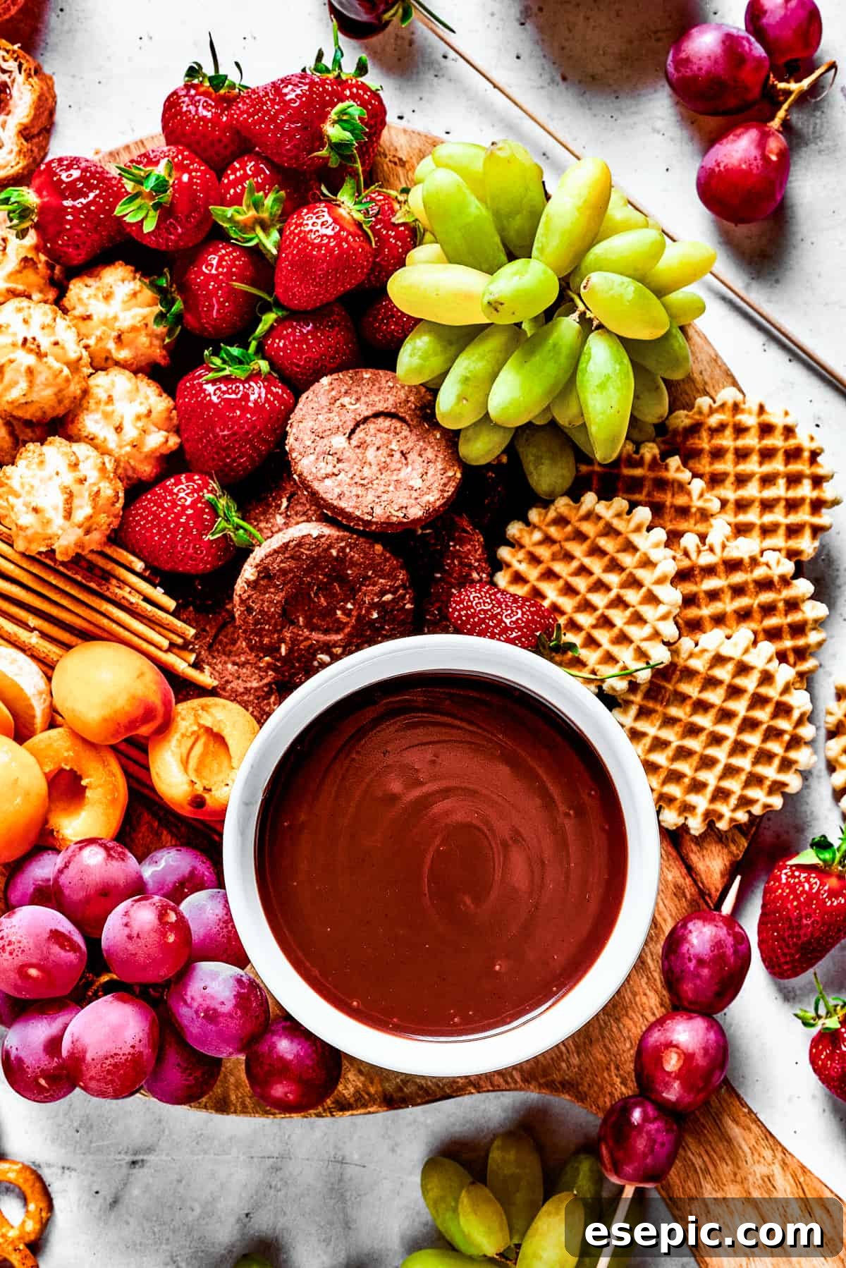 A beautifully arranged dessert board featuring various fruits, waffles, and pretzels, with a central bowl of rich chocolate fondue, inviting guests to partake.