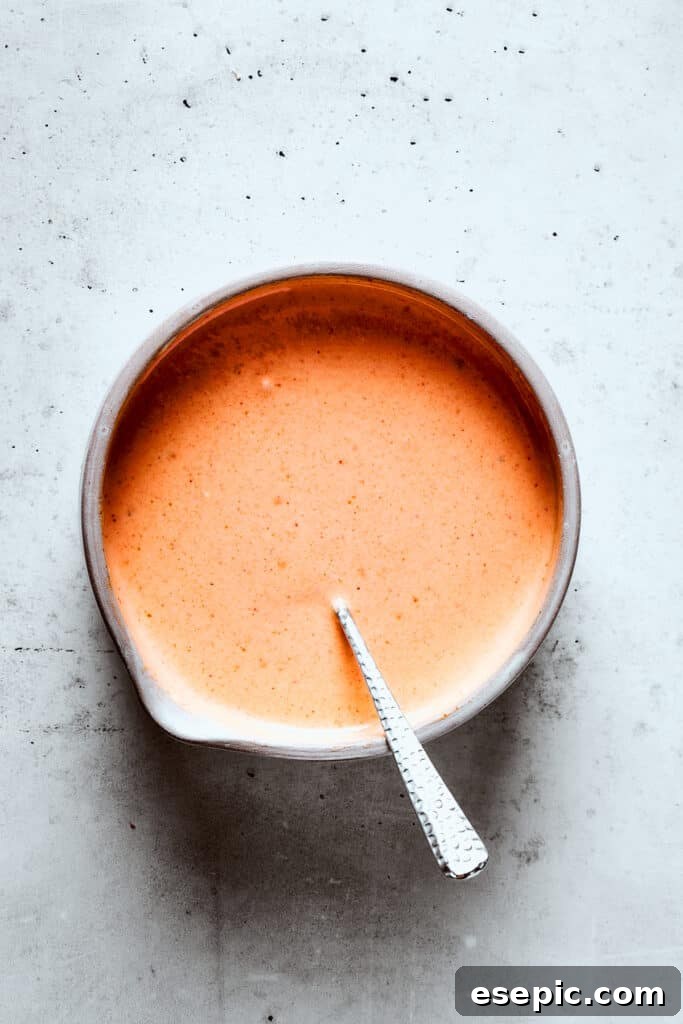 A hand mixing the Yum Yum Sauce ingredients in a glass bowl with a spoon, showing the creamy texture forming.