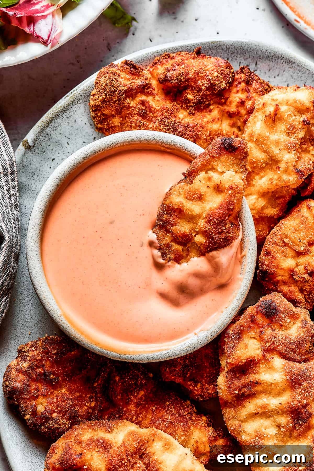 A close-up of a crispy chicken strip being dipped into a small bowl of homemade Yum Yum Sauce, with other chicken strips on a platter.