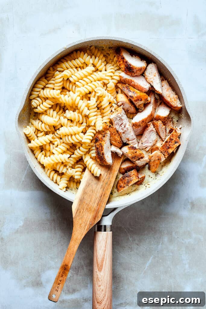 Cooked chicken strips and al dente pasta being gently tossed into the creamy ranch sauce in a skillet.
