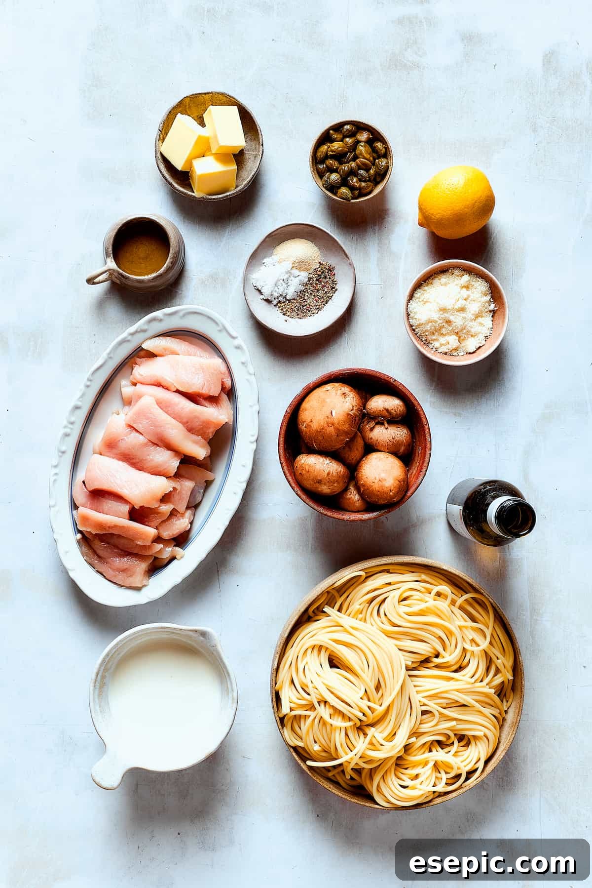 A collection of fresh ingredients for a homemade chicken piccata, including chicken breasts, mushrooms, lemons, capers, cream, and herbs, neatly arranged on a kitchen counter.