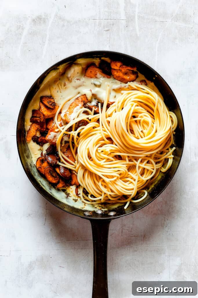 Adding cooked mushrooms, chicken, and pasta into the rich, creamy sauce in a skillet, ready to be combined.