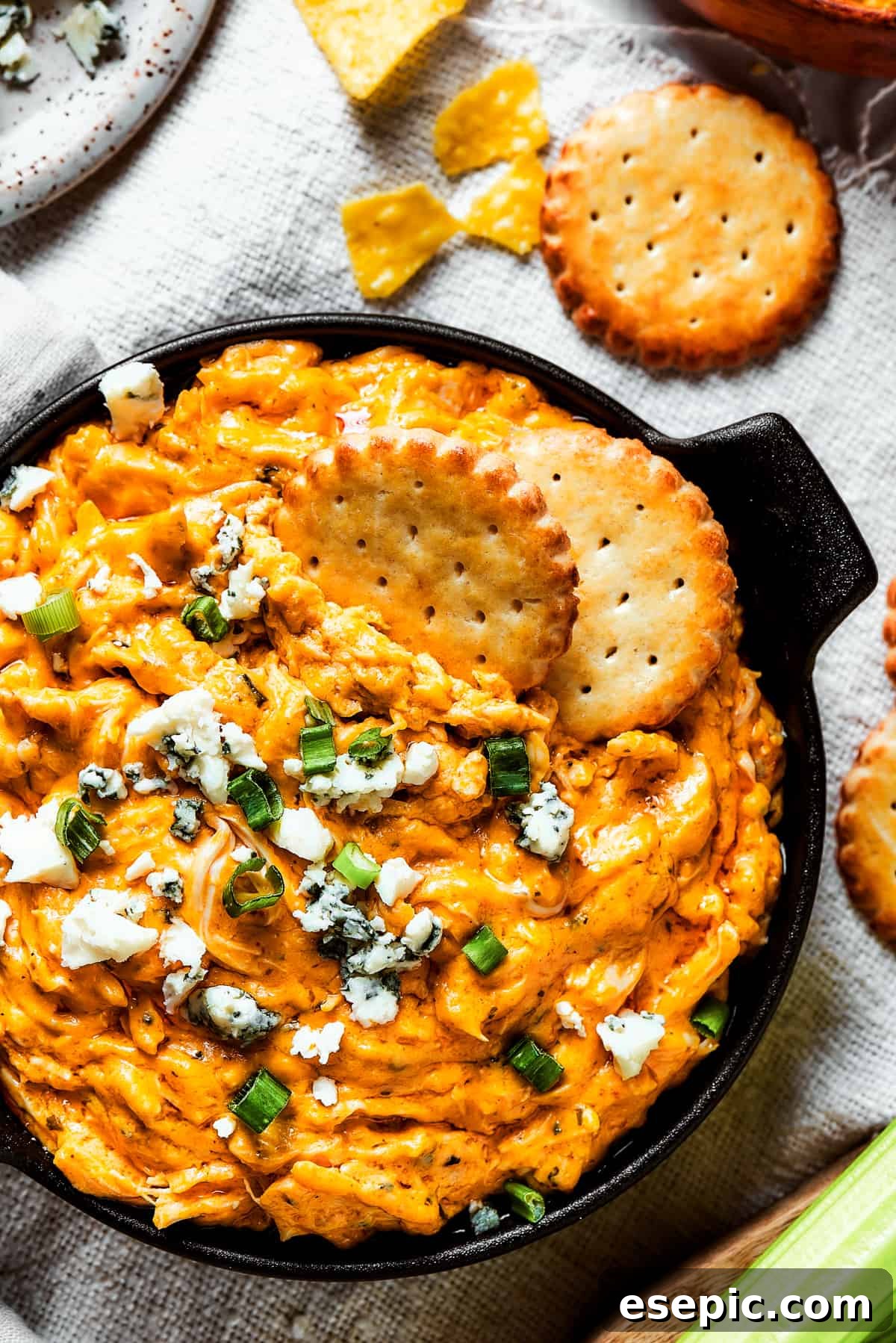 With its creamy, cheesy, spicy goodness, Crockpot Buffalo Chicken Dip is a favorite for parties, game days, movie night, and more! Overhead shot of a vibrant Crockpot Buffalo Chicken Dip, garnished with fresh green onions and crumbled blue cheese, served with crisp crackers and fresh celery sticks for dipping. The colors are rich and inviting, promising a delicious experience.