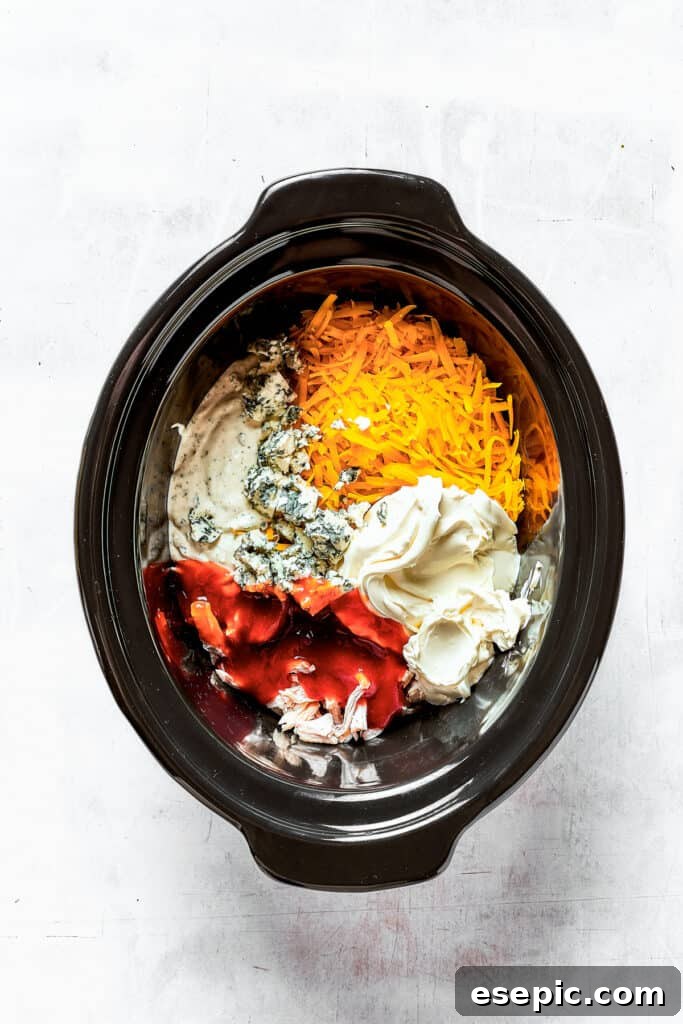 With its creamy, cheesy, spicy goodness, Crockpot Buffalo Chicken Dip is a favorite for parties, game days, movie night, and more! A tantalizing close-up of cream cheese, blue cheese, and tender shredded chicken being gently mixed with other vibrant ingredients inside a slow cooker insert, capturing the rich textures and promising flavors before cooking.