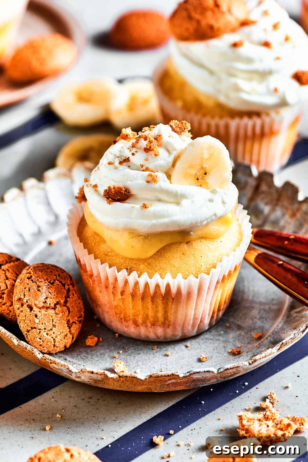 Decadent Banana Pudding Cupcakes 17 A single, meticulously prepared banana pudding cupcake rests on a white dessert plate, with a soft-focus background of more cupcakes awaiting enjoyment.