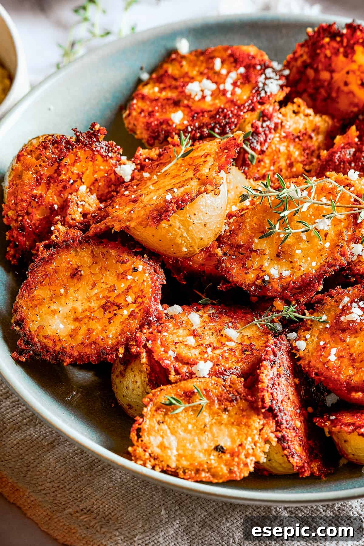 Cheesy roasted potatoes in a bowl next to cups of dipping sauce, a cloth napkin, and a serving on a plate, highlighting presentation and versatility.