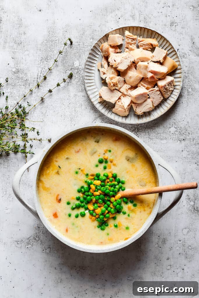 Adding chicken, peas, and corn to the pot pie soup. Cooked chicken, frozen peas, and corn being added to a bubbling pot of creamy chicken pot pie soup, with fresh thyme sprigs on the side.