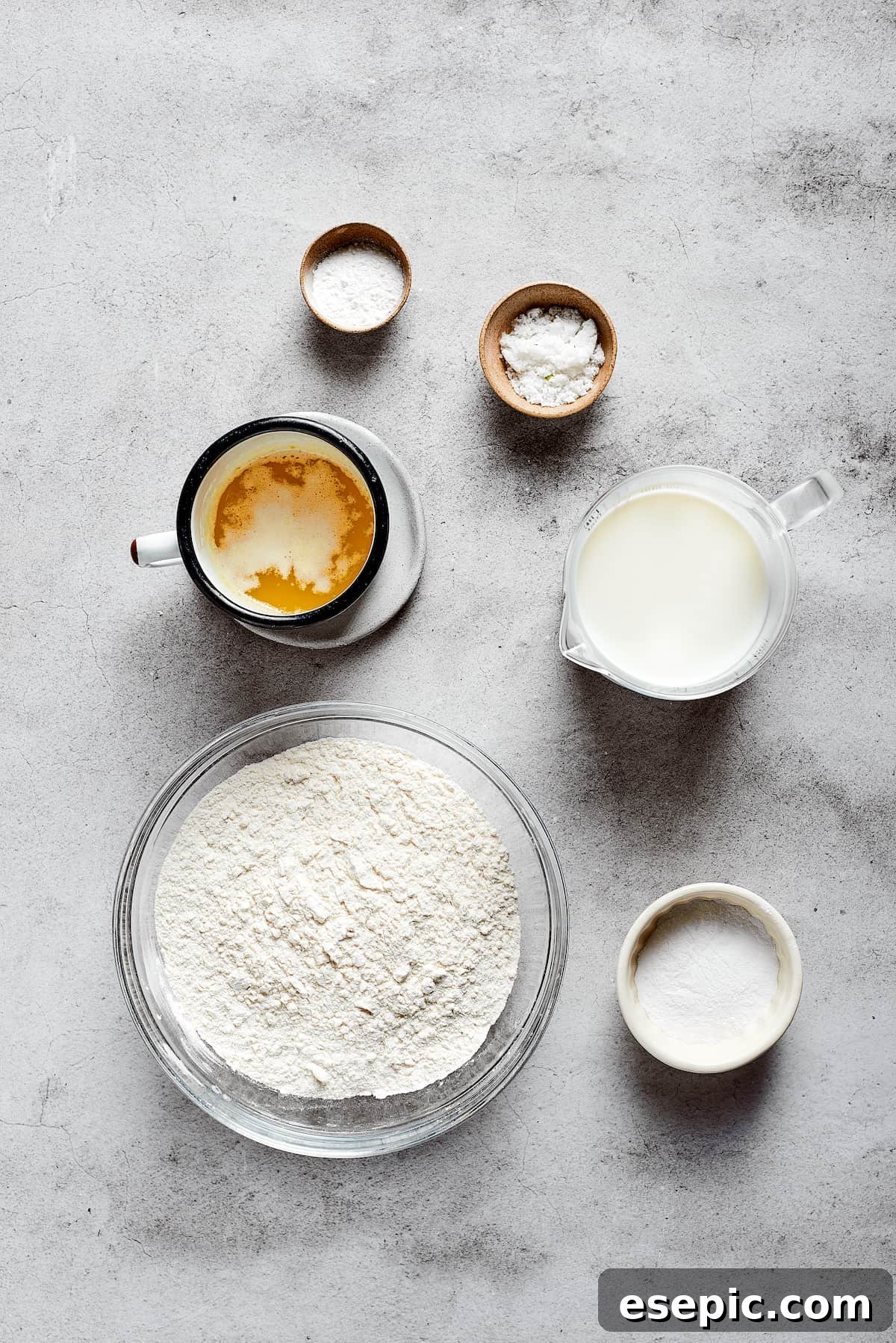 Overhead view of the ingredients needed for butter swim biscuits: a bowl of flour, a cup of melted butter, a pyrex of buttermilk, a bowl of sugar, a bowl of salt, and a bowl of baking powder, all neatly arranged.