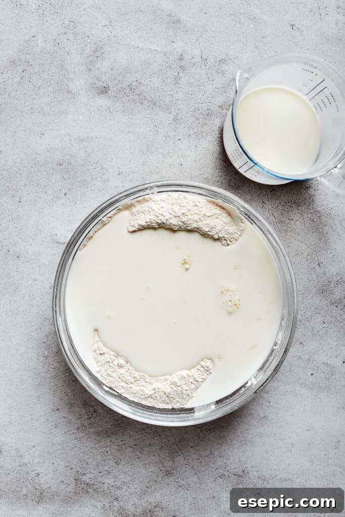 A mixing bowl with a pool of buttermilk on top of flour, next to a glass of buttermilk, illustrating the addition of liquid.