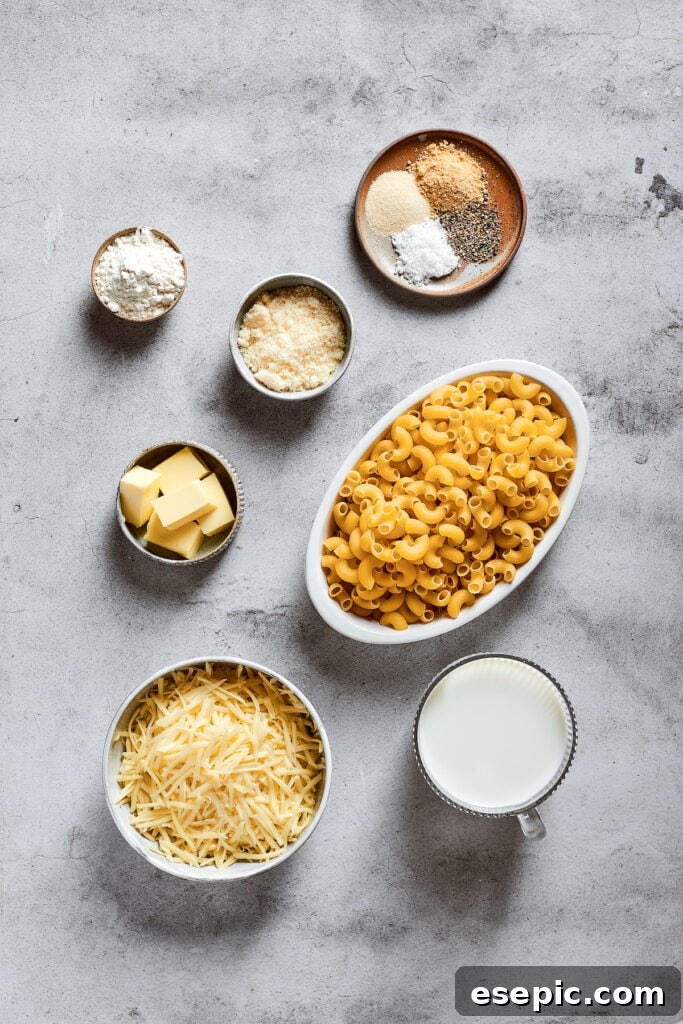 Overhead view of the ingredients needed for mac and cheese: a bowl of elbow macaroni, a bowl of shredded mozzarella cheese, a bowl of grated parmesan, a glass of milk, a bowl of flour, a bowl of butter, and a bowl with salt, pepper, garlic powder, and onion powder.