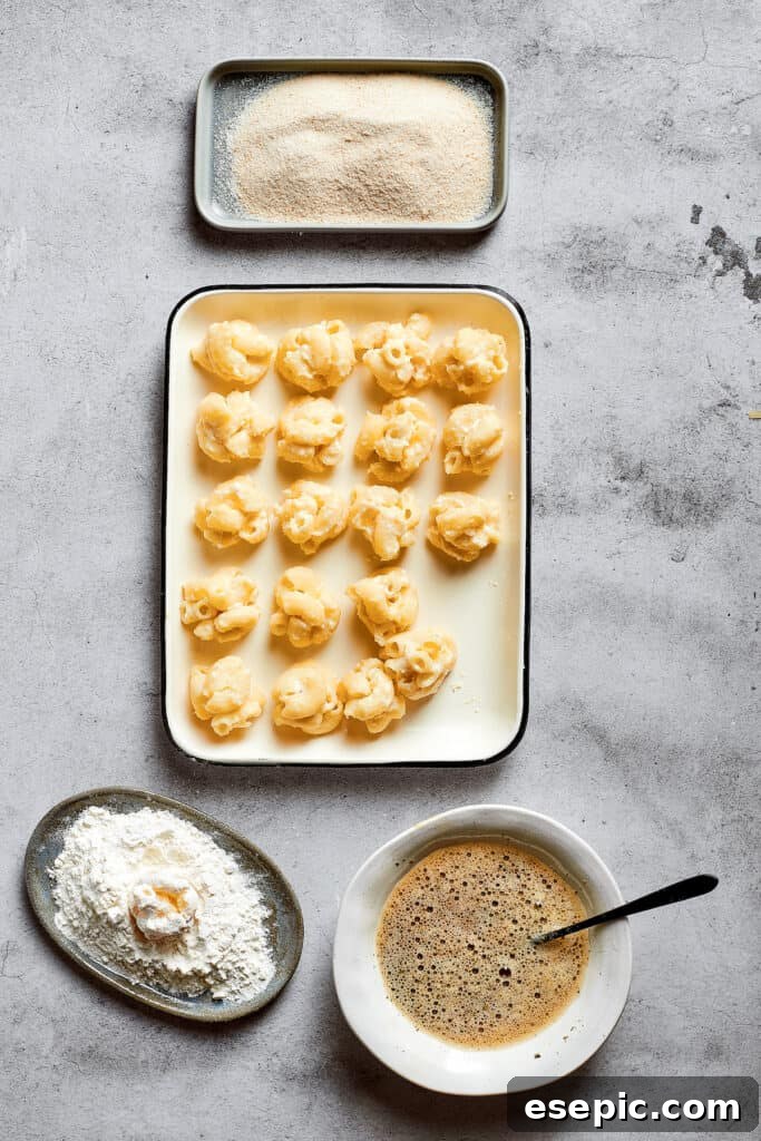 Overhead view of a tray of rolled balls of mac and cheese, next to a bowl of flour, a bowl of breadcrumbs, and a bowl of eggs beaten with Italian seasoning.