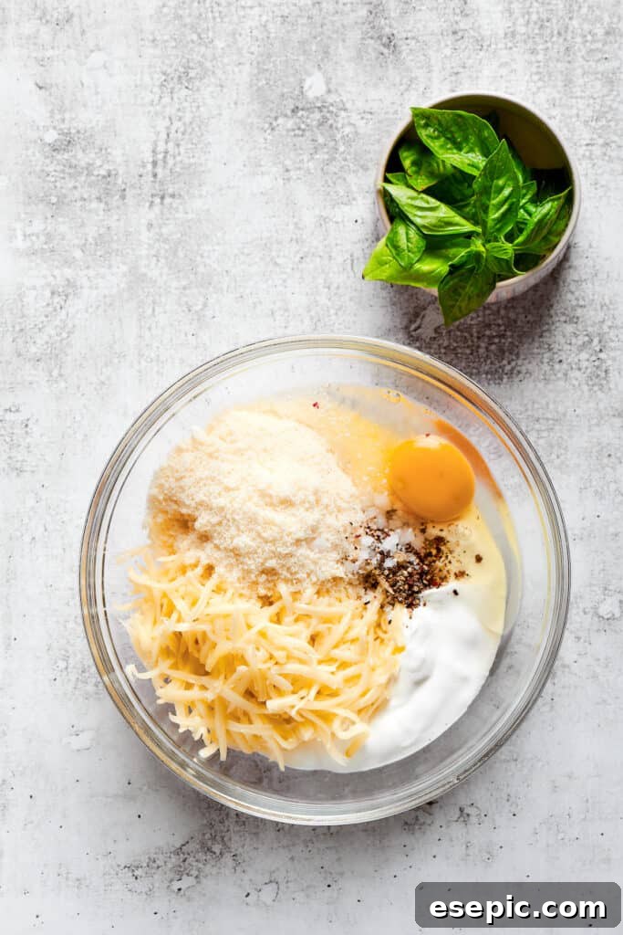 Overhead view of a mixing bowl filled with parmesan cheese, mozzarella cheese, ricotta, an egg, salt, and pepper, next to a bowl of basil.