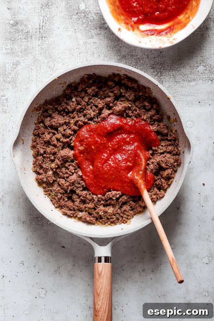 Overhead view of marinara sauce on top of cooked ground beef in a skillet with a wooden spoon.