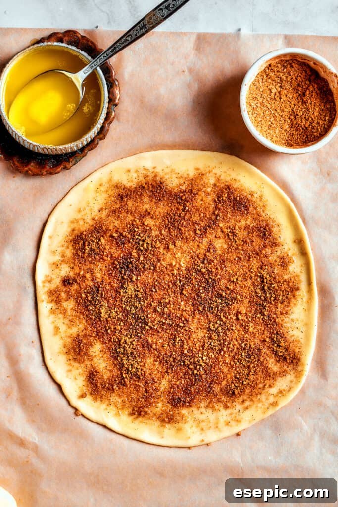 Overhead view of a piece of dough rolled into a circle, covered in cinnamon-sugar, next to a bowl of butter and a bowl of cinnamon-sugar.