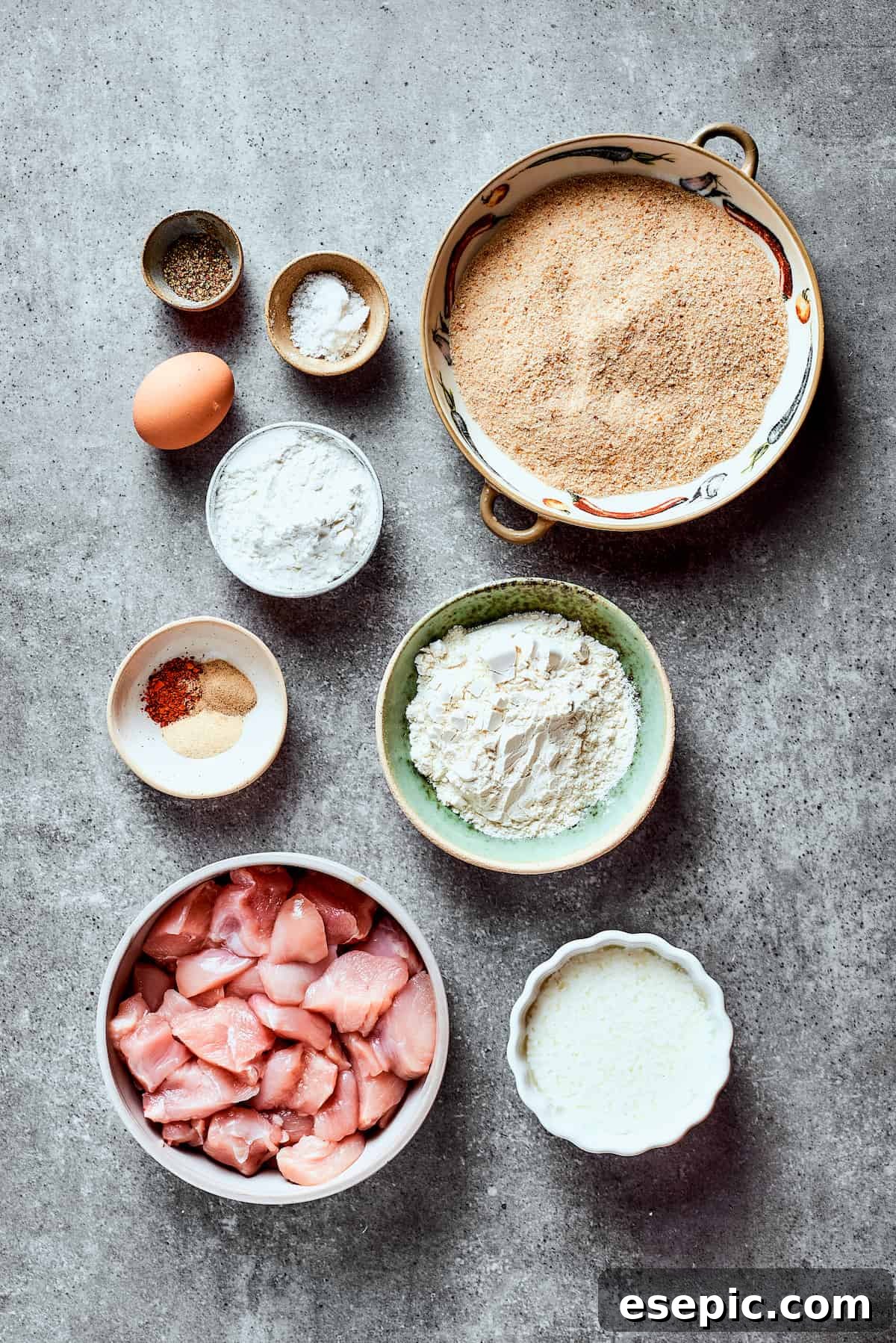 Overhead view of the ingredients needed for bang bang chicken: a bowl of chicken slices, a bowl of flour, a bowl of panko breadcrumbs, a bowl of buttermilk, a bowl of cornstarch, an egg, a bowl of salt, a bowl of pepper, and a bowl of garlic powder, chili powder, and white pepper.