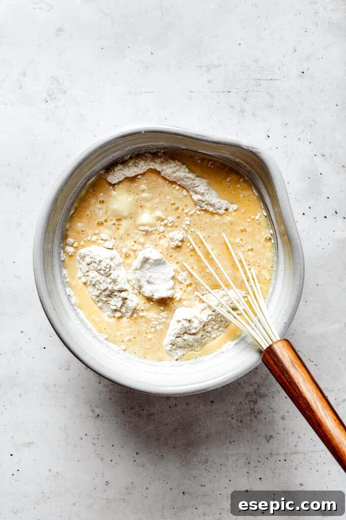 Overhead view of a mixing bowl with wet ingredients, flour, and a whisk.