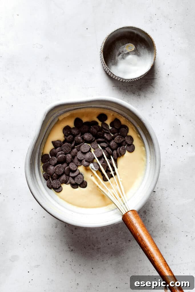 Overhead view of chocolate chips on top of pancake batter in a mixing bowl with a whisk, next to an empty bowl.