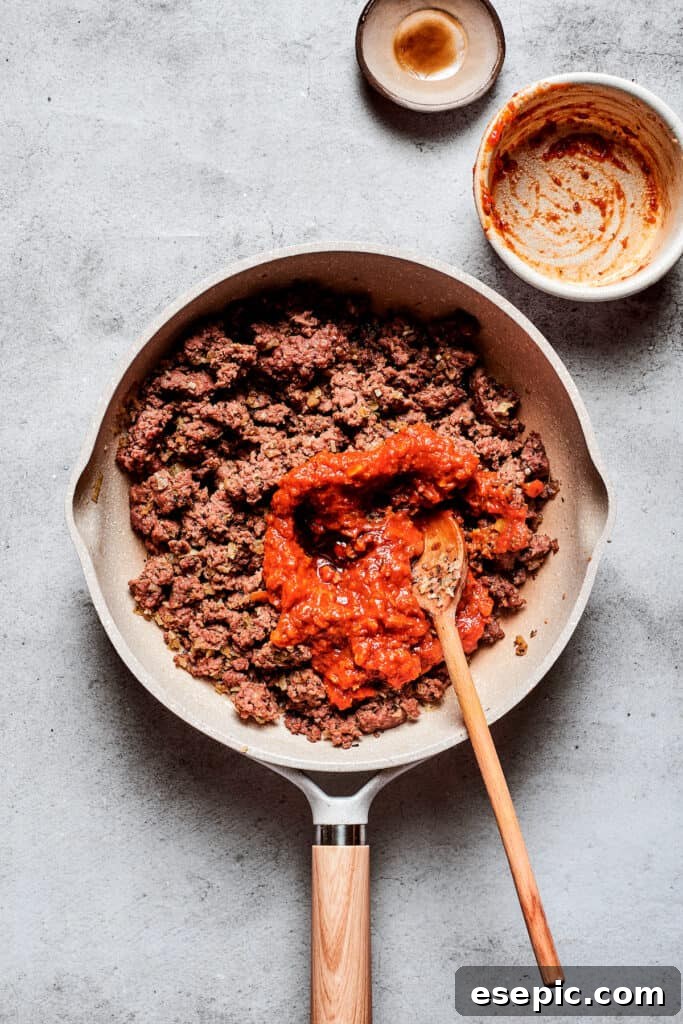 A vibrant pizza sauce is generously stirred into a pan of perfectly browned ground meat, infusing it with rich, savory flavor.