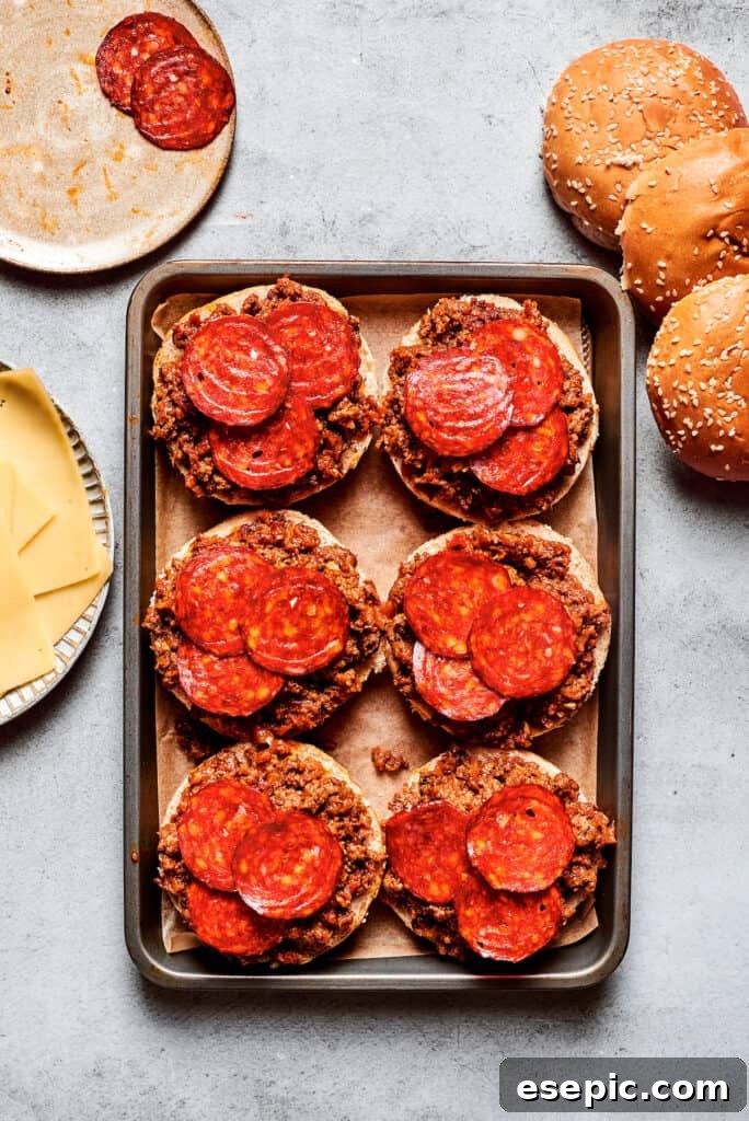 Crisp pepperoni slices are artfully placed on top of savory pizza burgers on a tray, adding a classic touch before the cheese.