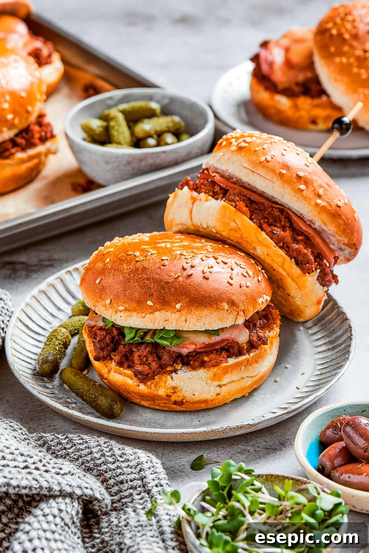 Two perfectly constructed pizza burgers, garnished with fresh pickles, are served on a plate, with additional burgers and sides blurred in the background, hinting at a larger, delicious spread.